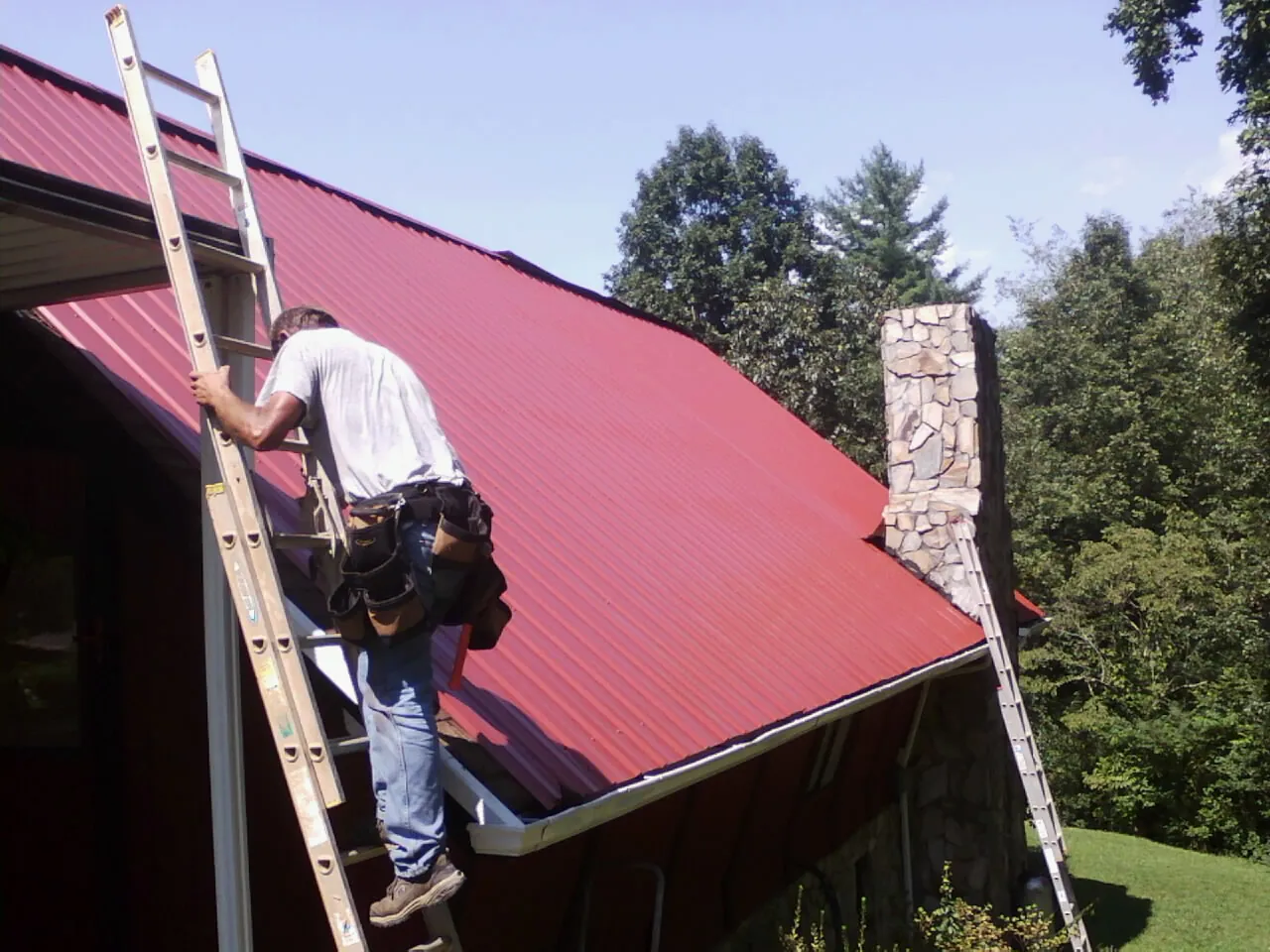 Roofer on a ladder beside a red metal roof and stone chimney — Soffit & Fascia Repair by Roofing Brothers in Ruffs Dale